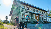 Der Berggasthof Dreisesse im Bayerischen Wald steht im Hintergrund von zwei Wanderern mit Hund.