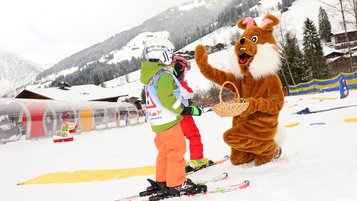 Kind auf Skiern gibt einem Hasen-Maskottchen mit Korb auf der Skipiste ein High-Five, im Hintergrund verschneite Berge und Skihütten.