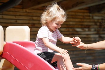 Kleines Kind rutscht lachend eine rote Rutsche hinunter und wird von einer Betreuerin im Familienhotel Sonne Bezau aufgefangen.