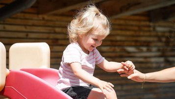 Kleines Kind rutscht lachend eine rote Rutsche hinunter und wird von einer Betreuerin im Familienhotel Sonne Bezau aufgefangen.