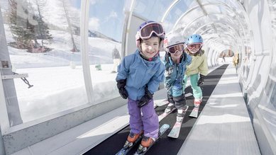 Drei Kinder fahren mit Skiern auf einem überdachten Zauberteppich im Winterurlaub beim Familienhotel Gorfion in Liechtenstein.