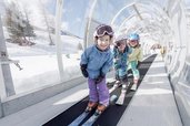 Drei Kinder fahren mit Skiern auf einem überdachten Zauberteppich im Winterurlaub beim Familienhotel Gorfion in Liechtenstein.