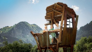 Kinder stehen lachend auf einem hölzernen Aussichtsturm mit Bergblick am Waldspielplatz des Familienhotels Krone im Allgäu.