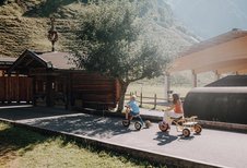 Kinder fahren im Außenbereich des Familienhotels Oberkarteis mit kleinen Fahrzeugen auf einem Weg nahe eines Holzgebäudes.