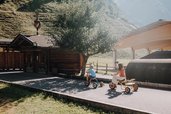 Kinder fahren im Außenbereich des Familienhotels Oberkarteis mit kleinen Fahrzeugen auf einem Weg nahe eines Holzgebäudes.