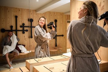 Frau im Bademantel föhnt sich die Haare im Wellnessbereich des Zauchenseehof, während ein Mann im Hintergrund entspannt ein Handtuch nutzt; warmes Holzdesign schafft eine gemütliche Atmosphäre.
