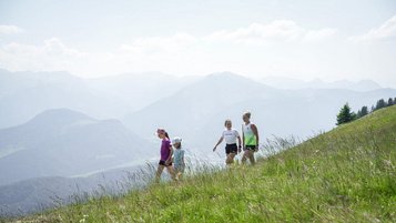 Familie wandert über eine grüne Bergwiese mit weitem Blick auf die Alpen beim Familienhotel Sonne Bezau.