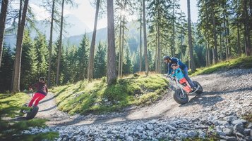 Ein Vater und sein Kind fahren gemeinsam mit einem Monsterroller eine kurvige Schotterstrecke hinunter, während eine Frau vor ihnen elegant die Kurve nimmt.