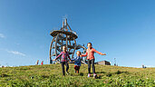 Wandern in der Rhön: Kinder laufen über eine Wiese im Hintergrund der Aussichtsturm auf dem Naturlehrpfad "Ellenbogen"