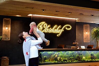 Mutter hält ihr Baby mit den Händen in die Höhe vor der Rezeption des Familienhotels Ulrichshof im Bayerischen Wald.